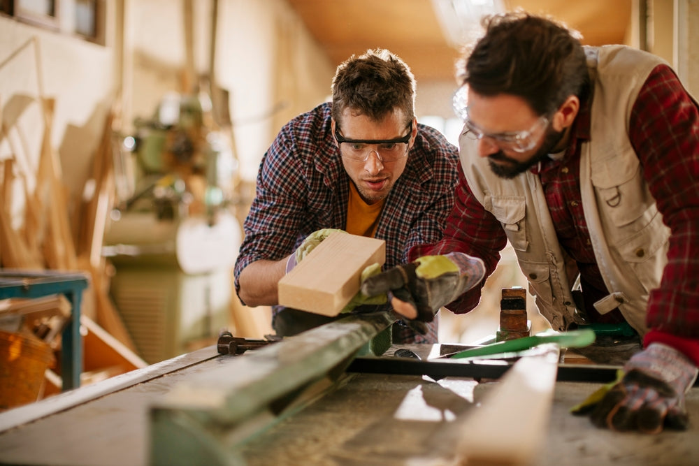Two carpenters working together in a woodworking shop wearing safety gear